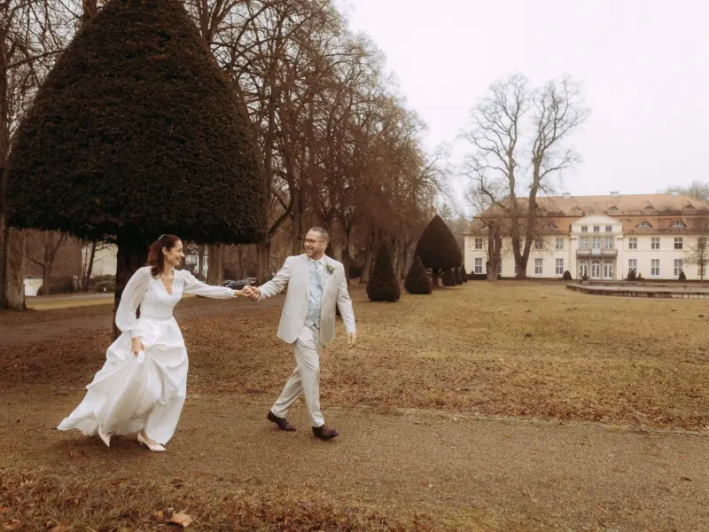 Ein glückliches Brautpaar hält Händchen und spaziert über einen Kiesweg vor einem herrschaftlichen Schloss in einer herbstlichen Parklandschaft. Die Braut trägt ein elegantes weißes Kleid, der Bräutigam einen hellen Anzug. Eine stimmungsvolle Aufnahme vom Hochzeitsfotograf in Schwerin und MV.