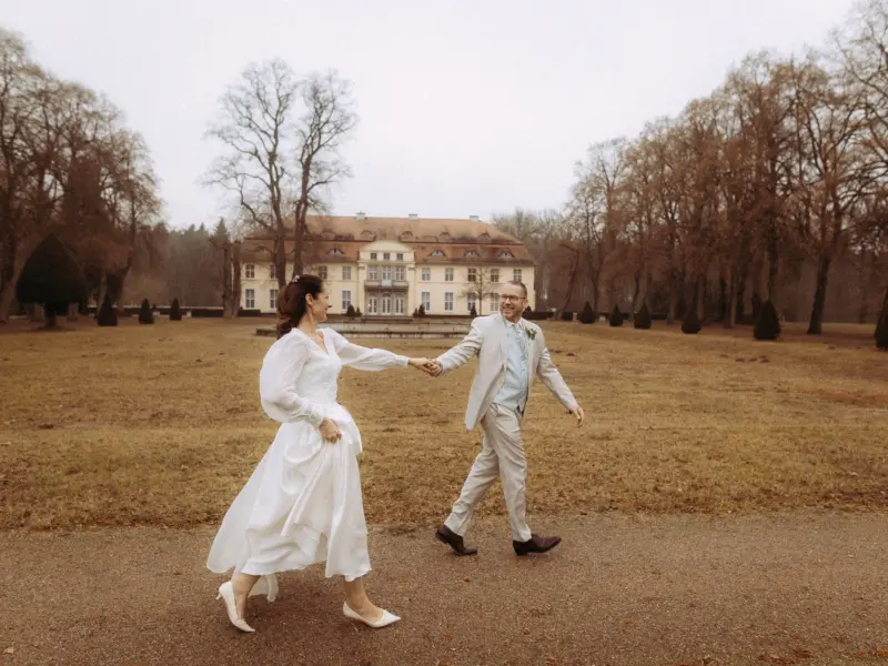 Ein glückliches Brautpaar hält Händchen und spaziert vor einem herrschaftlichen Schloss in einer herbstlichen Parkanlage. Die Braut trägt ein weißes Hochzeitskleid, der Bräutigam einen hellen Anzug. Eine stimmungsvolle Aufnahme von deinem Hochzeitsfotograf in Schwerin und MV.