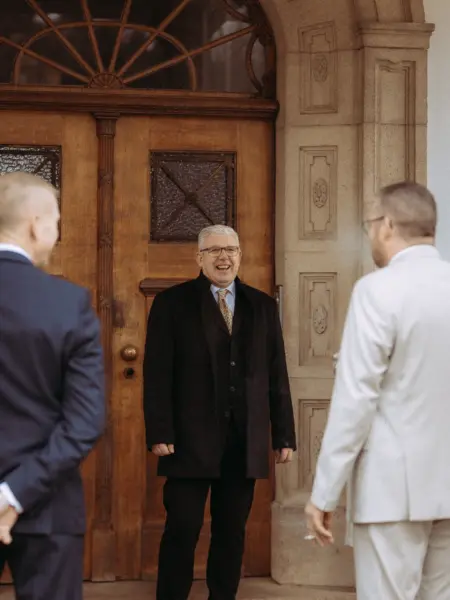 Ein fröhlich lachender Mann in einem eleganten schwarzen Mantel und Anzug steht vor einer historischen Holztür, während zwei Männer in Anzügen im Vordergrund stehen – eine Szene, die ein erfahrener Hochzeitsfotograf in Schwerin und MV festhält.
