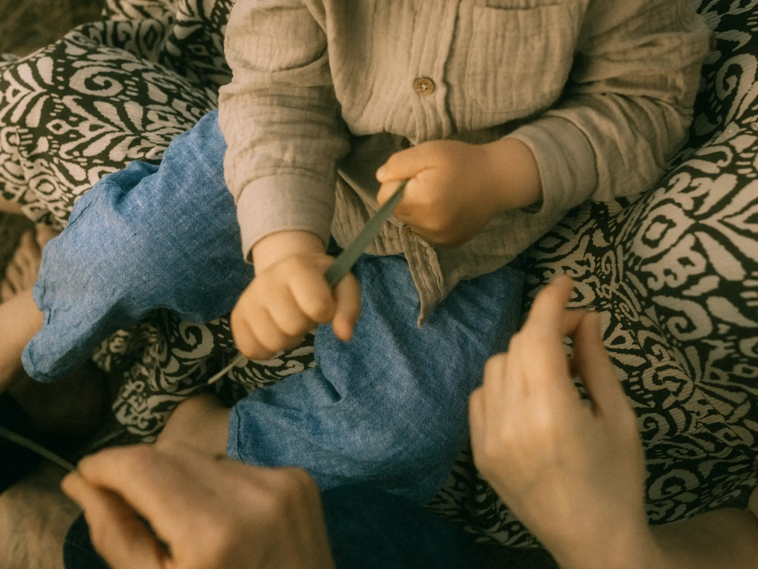 Ein Kleinkind in einem beigen Hemd und blauen Jeans spielt konzentriert mit einem langen Grashalm, während die Hände der Eltern liebevoll unterstützend daneben zu sehen sind. Eine warme, intime Aufnahme, wie sie ein Familienfotograf in Schwerin festhält.