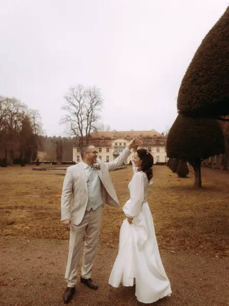 Ein Brautpaar tanzt glücklich vor einem historischen Schloss im herbstlichen Garten. Der Bräutigam im beigen Anzug führt die Braut in ihrem eleganten weißen Hochzeitskleid. Professionelle Hochzeitsfotografie von einem Hochzeitsfotograf in Schwerin und MV bei einer romantischen Hochzeit.