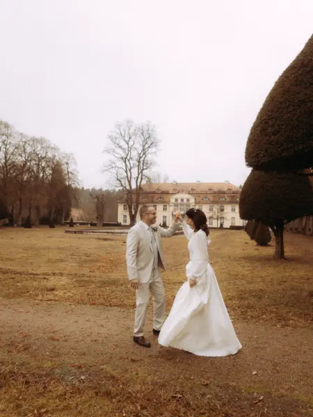 Ein Brautpaar tanzt glücklich vor einem herrschaftlichen Schloss in einer herbstlichen Parkanlage. Die Braut trägt ein elegantes weißes Kleid, der Bräutigam einen beigen Anzug. Diese romantische Aufnahme entstand durch einen professionellen Hochzeitsfotograf in Schwerin und MV.