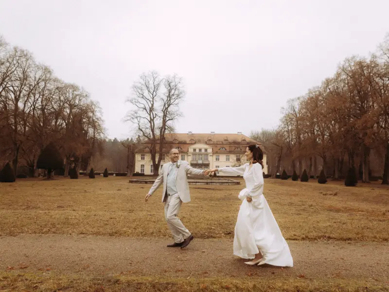 Ein glückliches Brautpaar tanzt Hand in Hand auf einem Kiesweg vor einem historischen Schloss in einer herbstlichen Parkanlage. Die Szene fängt einen romantischen Moment ein, ideal für eine Lovestory als Fotograf für Pärchen in Schwerin.