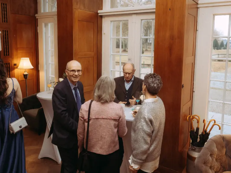 Eine Gruppe von Hochzeitsgästen unterhält sich in einem eleganten, holzgetäfelten Raum bei einem Empfang. Während ein Herr in den Anzug lächelt, stehen zwei Damen und ein weiterer Herr um einen Stehtisch. Dieser Moment wurde von einem Hochzeitsfotograf in Schwerin und MV festgehalten.