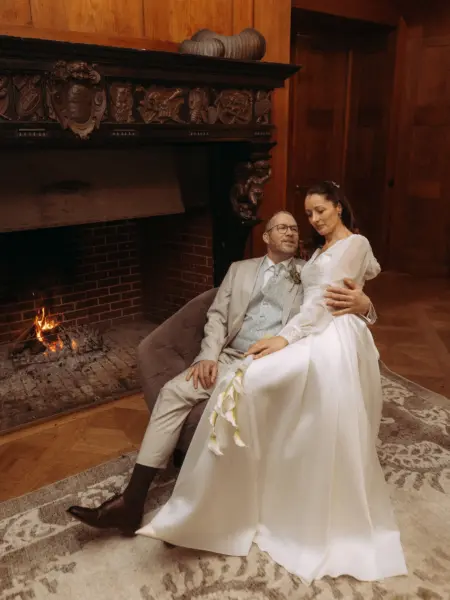 Ein Brautpaar in eleganter Hochzeitskleidung sitzt in einem historischen Raum vor einem prasselnden Kaminfeuer. Die Braut hält eine Callablüte, während der Bräutigam sie liebevoll umfasst. Eine stimmungsvolle Aufnahme vom Lovestory Fotograf in Schwerin und MV.