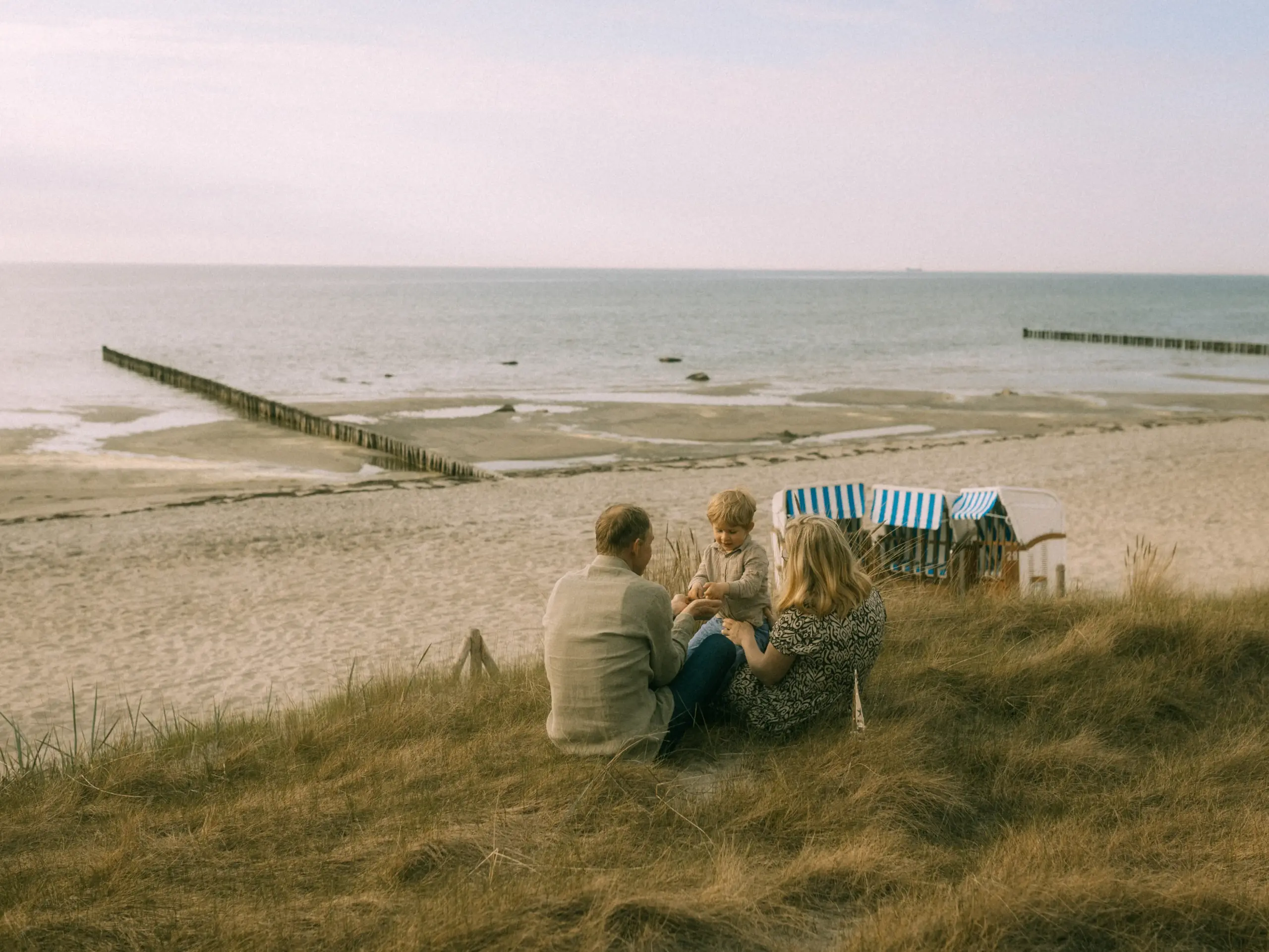 Eine junge Familie sitzt in den Dünen an der Ostseeküste und genießt die gemeinsame Zeit. Im Hintergrund sind der Strand und traditionelle Strandkörbe zu sehen. Authentische Familienfotos in Schwerin und Umgebung festgehalten für bleibende Erinnerungen.