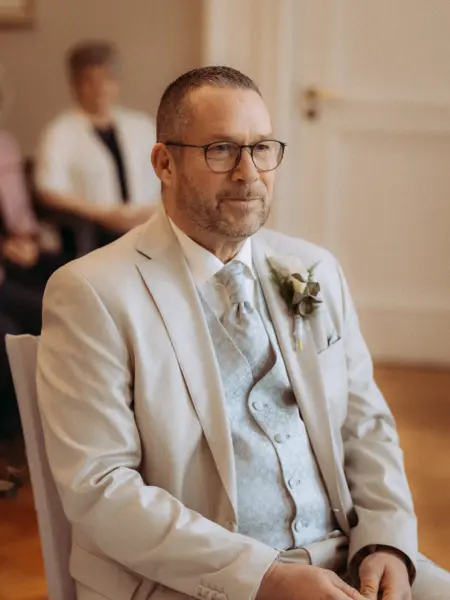 Ein Bräutigam mit Brille und Bart trägt einen eleganten beigen Hochzeitsanzug mit gemusterter Weste und Krawatte. Er sitzt ruhig bei der Zeremonie, während im Hintergrund Gäste unscharf zu sehen sind. Professionell festgehalten von Ihrem Hochzeitsfotograf in Schwerin und MV.