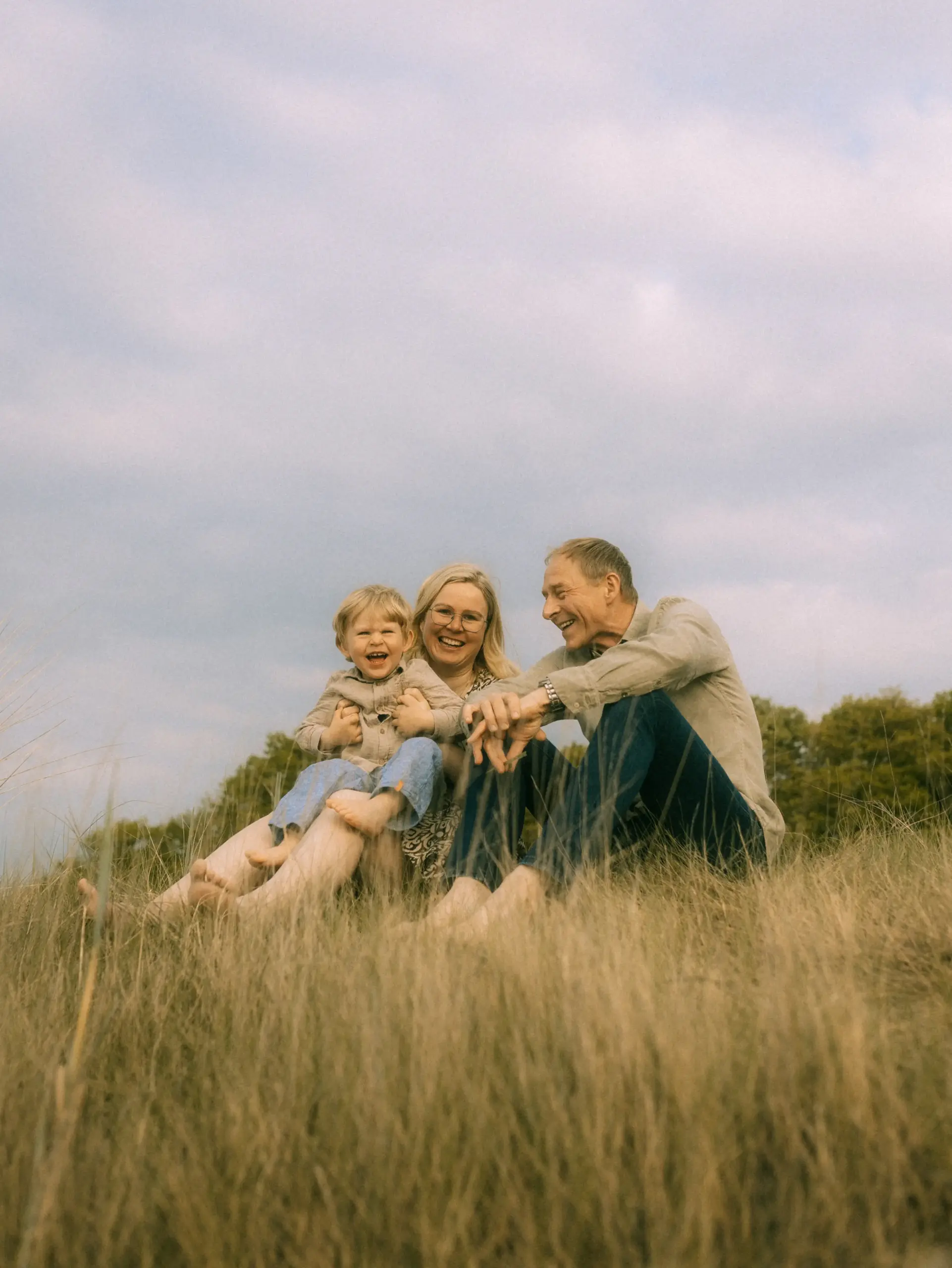 Ein glückliches Paar mit ihrem kleinen Sohn sitzt lachend im hohen Gras einer weiten Wiesenlandschaft. Die Familie genießt einen entspannten Moment in der Natur, eingefangen von einem professionellen Familienfotograf in Schwerin, der authentische Familienfotos in Schwerin erstellt.