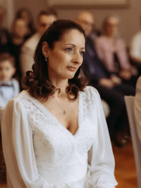 Eine elegante Braut mit brünettem Haar und weißem Spitzenkleid blickt bei einer Hochzeitszeremonie gefühlvoll zur Seite. Das stimmungsvolle Porträt wurde von einem Hochzeitsfotograf in Schwerin und MV festgehalten, während im Hintergrund verschwommene Gäste zu sehen sind.