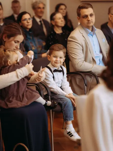 Ein kleiner Junge mit Fliege und Hosenträgern lächelt charmant in die Kamera, während er neben seiner Mutter und einem Kleinkind in einem festlichen Saal sitzt. Als Fotograf für Familien in Schwerin fange ich solche natürlichen und emotionalen Momente bei Feierlichkeiten ein.