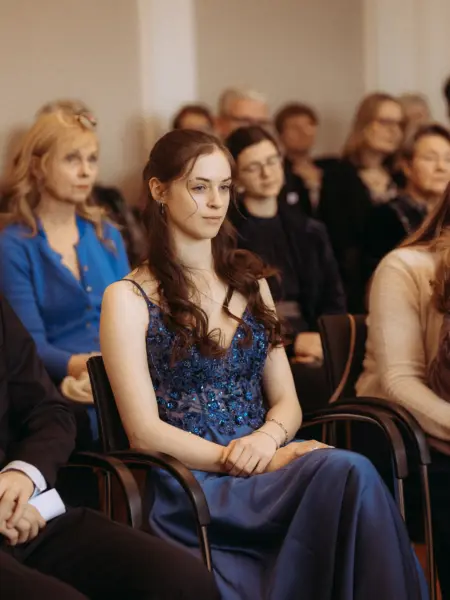 Eine junge Frau mit langem braunem Haar trägt ein elegantes, dunkelblaues Abendkleid mit Pailletten und sitzt aufmerksam bei einer Veranstaltung. Im Hintergrund sind weitere Gäste in einem hellen Raum zu sehen, eingefangen durch einen professionellen Hochzeitsfotograf in Schwerin und MV.