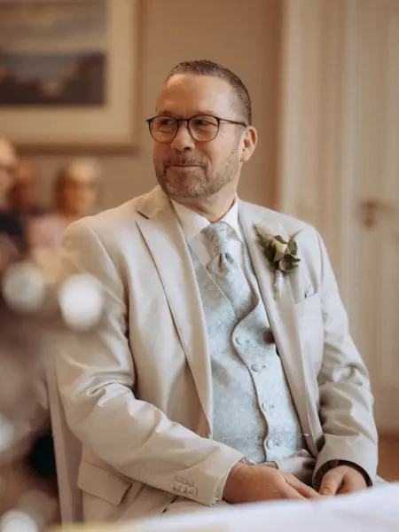 Ein Bräutigam in einem eleganten hellen Hochzeitsanzug mit passender gemusterter Weste und Ansteckblume blickt lächelnd bei einer Zeremonie zur Seite. Professionelle Aufnahme von einem Hochzeitsfotograf in Schwerin und MV, die eine emotionale und feierliche Stimmung einfängt.