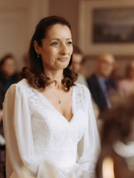 Eine elegante Braut mit braunem Haar trägt ein weißes Spitzenkleid mit langen Ärmeln und blickt während der Trauung mit einem sanften Lächeln in die Ferne. Diese Aufnahme wurde von einem Hochzeitsfotograf in Schwerin und MV festgehalten.
