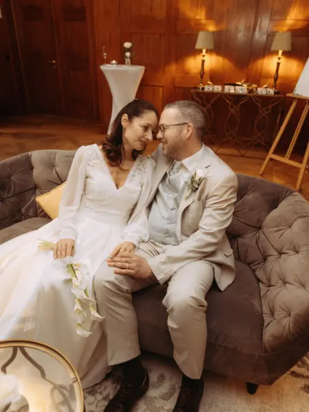 Ein Brautpaar sitzt sich liebevoll anblickend auf einem eleganten grauen Samtsofa in einem festlich dekorierten Raum. Die Braut trägt ein weißes Kleid mit langen Ärmeln und hält Callas, während der Bräutigam einen hellen Anzug trägt. Ein Hochzeitsfotograf in Schwerin und MV.