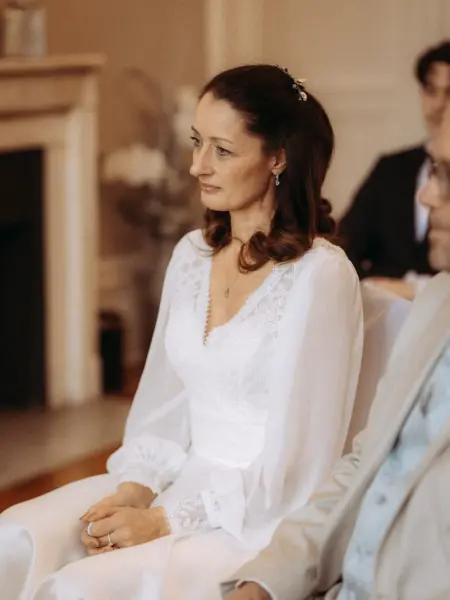Eine Braut mit elegantem, langärmeligem Spitzenkleid sitzt während einer Hochzeitszeremonie andächtig und lächelnd in einem hellen Raum. Dieses stimmungsvolle Porträt wurde von einem Hochzeitsfotograf in Schwerin und MV aufgenommen.