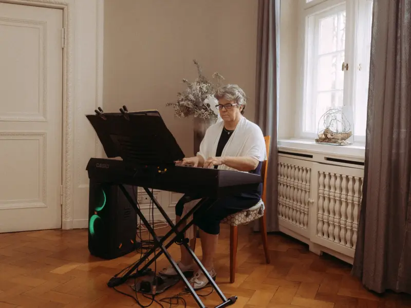 Eine ältere Dame mit Brille und weißem Überwurf spielt konzentriert auf einem Casio-Digitalpiano in einem hellen, stilvoll eingerichteten Raum. Neben ihr steht ein leuchtender JBL-Lautsprecher. Die Aufnahme entstand durch einen professionellen Hochzeitsfotograf in Schwerin und MV.