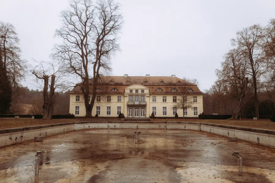Ein herrschaftliches, hellgelbes Schloss mit symmetrischer Architektur und Walmdach hinter einem leeren, reflektierenden Wasserbecken an einem bewölkten Tag. Die Szenerie mit kahlen Bäumen bietet eine elegante Kulisse für einen Hochzeitsfotograf in Schwerin und MV.