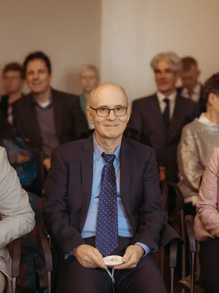 Ein freundlich lächelnder, älterer Herr mit Brille und dunkelblauem Anzug sitzt bei einer Hochzeitszeremonie und hält behutsam die Eheringe in seinen Händen. Im Hintergrund sind weitere Gäste zu sehen. Professionelle Aufnahme durch Ihren Hochzeitsfotograf in Schwerin und MV.