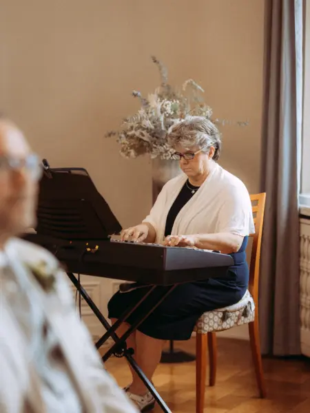 Eine ältere Dame mit Brille spielt konzentriert auf einem E-Piano während einer Hochzeitsfeier. Im Vordergrund ist unscharf eine Person zu sehen, im Hintergrund steht ein elegantes Blumenarrangement. Professionelle Aufnahme von Ihrem Hochzeitsfotograf in Schwerin und MV.