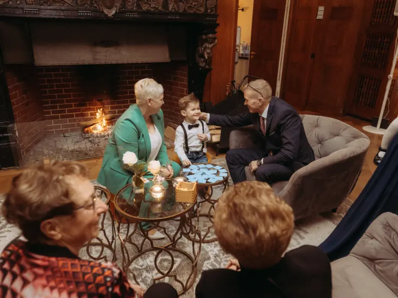Ein kleiner Junge mit Fliege und Hosenträgern sitzt zwischen seinen Großeltern vor einem gemütlichen Kamin, während sie gemeinsam spielen. Eine herzliche Szene, die die familiäre Verbundenheit bei einer Feier zeigt – eingefangen vom Familienfotograf in Schwerin.
