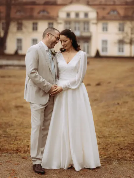Ein glückliches Brautpaar steht sich liebevoll gegenüber vor einem historischen Schloss, festgehalten von einem Hochzeitsfotograf in Schwerin und MV. Die Braut trägt ein elegantes weißes Kleid mit langen Ärmeln, der Bräutigam einen hellen Anzug, während sie sich zärtlich an den Händen halten.