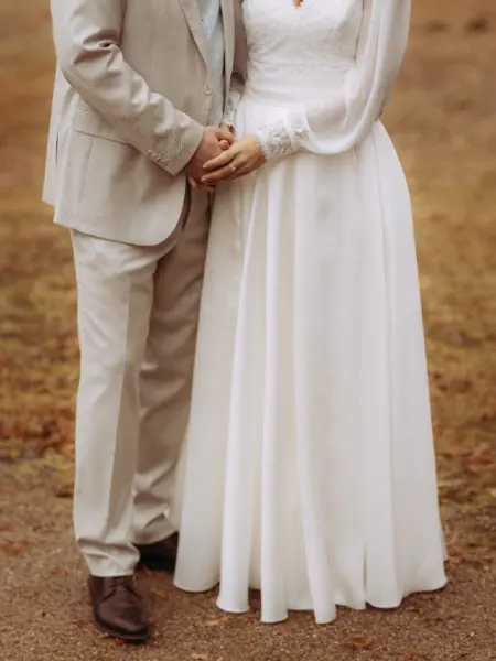 Ein Brautpaar hält sich liebevoll an den Händen. Die Braut trägt ein elegantes weißes Kleid mit Spitzenärmeln, der Bräutigam einen hellen Anzug. Die Aufnahme fängt einen intimen Moment im Freien ein, perfekt für eine Lovestory als Fotograf für Pärchen in Schwerin.