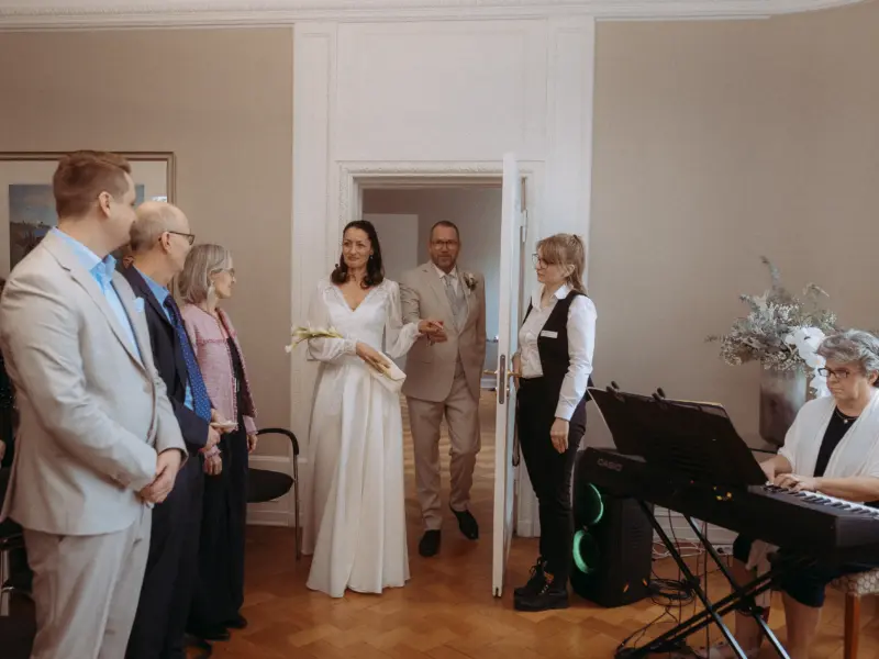 Eine Braut in einem eleganten weißen Kleid betritt händchenhaltend mit dem Bräutigam im hellen Anzug den Raum, während Gäste zuschauen und eine Musikerin am Keyboard spielt. Professionelle Momente eingefangen vom Hochzeitsfotograf in Schwerin und MV.