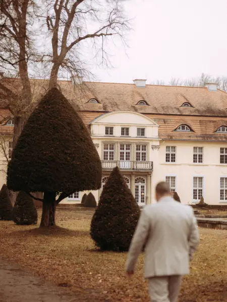 Ein Bräutigam im hellen Anzug geht durch den herbstlichen Park auf ein prachtvolles, historisches Schloss zu, aufgenommen von einem Hochzeitsfotograf in Schwerin und MV. Die Szene wirkt elegant und zeitlos vor der Kulisse des imposanten Gebäudes.