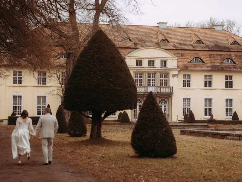 Ein Brautpaar spaziert händchenhaltend durch einen herbstlichen Park vor einem historischen Schloss. Die Braut trägt ein elegantes weißes Kleid, der Bräutigam einen hellen Anzug. Diese romantische Szene wurde von einem Lovestory Fotograf in Schwerin und MV festgehalten.