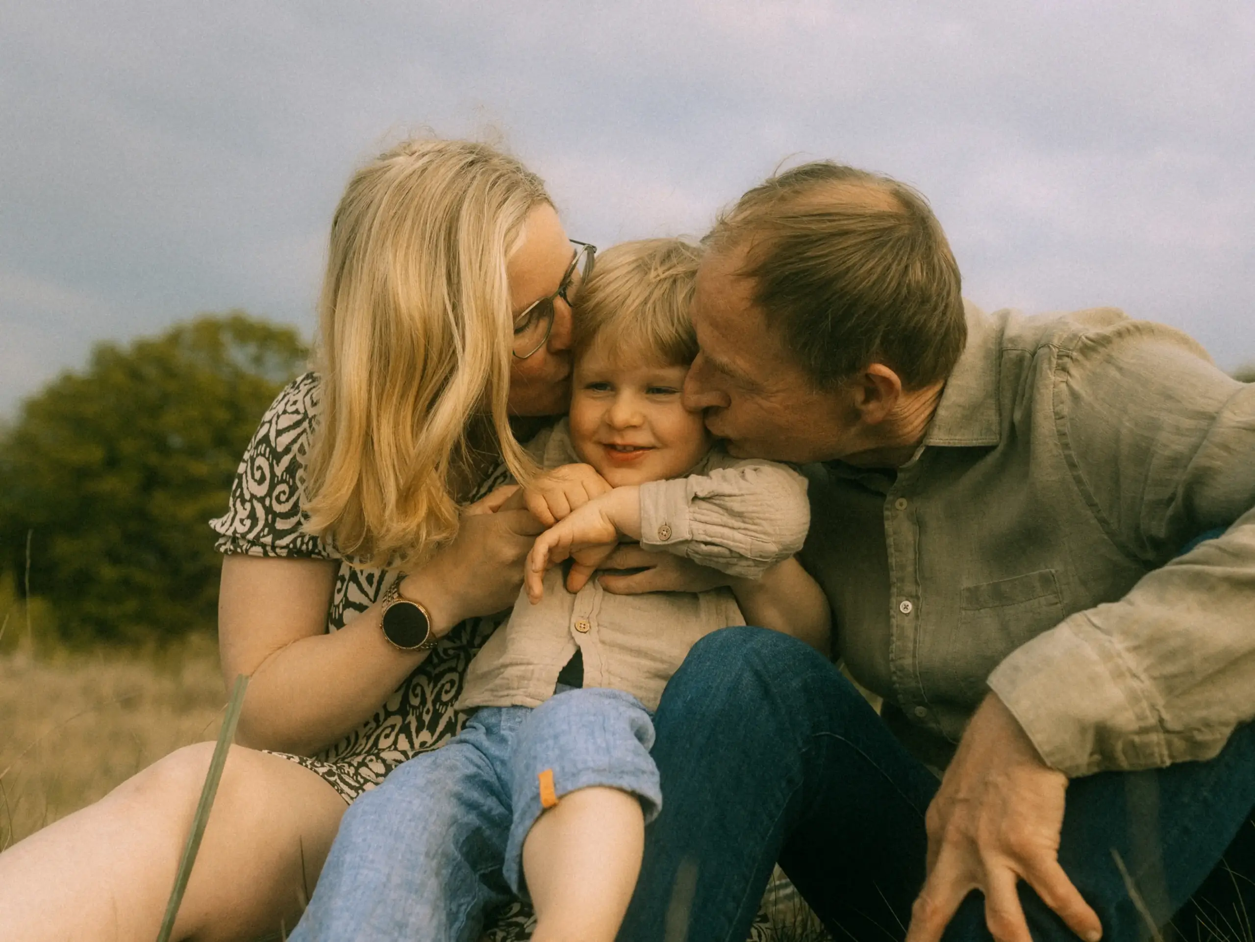 Ein glückliches Kleinkind sitzt zwischen seinen Eltern auf einer Wiese, während beide ihm liebevoll einen Kuss auf die Wange geben. Die warme, natürliche Atmosphäre unterstreicht die Verbundenheit dieser Familienfotos in Schwerin, die im Freien aufgenommen wurden.