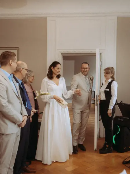Das Brautpaar betritt händchenhaltend einen eleganten Raum, begleitet von Gästen und einer Hochzeitsplanerin. Die Braut trägt ein weißes Langarmkleid und hält Callas, der Bräutigam einen hellen Anzug. Ein authentischer Moment, festgehalten vom Hochzeitsfotograf in Schwerin und MV.
