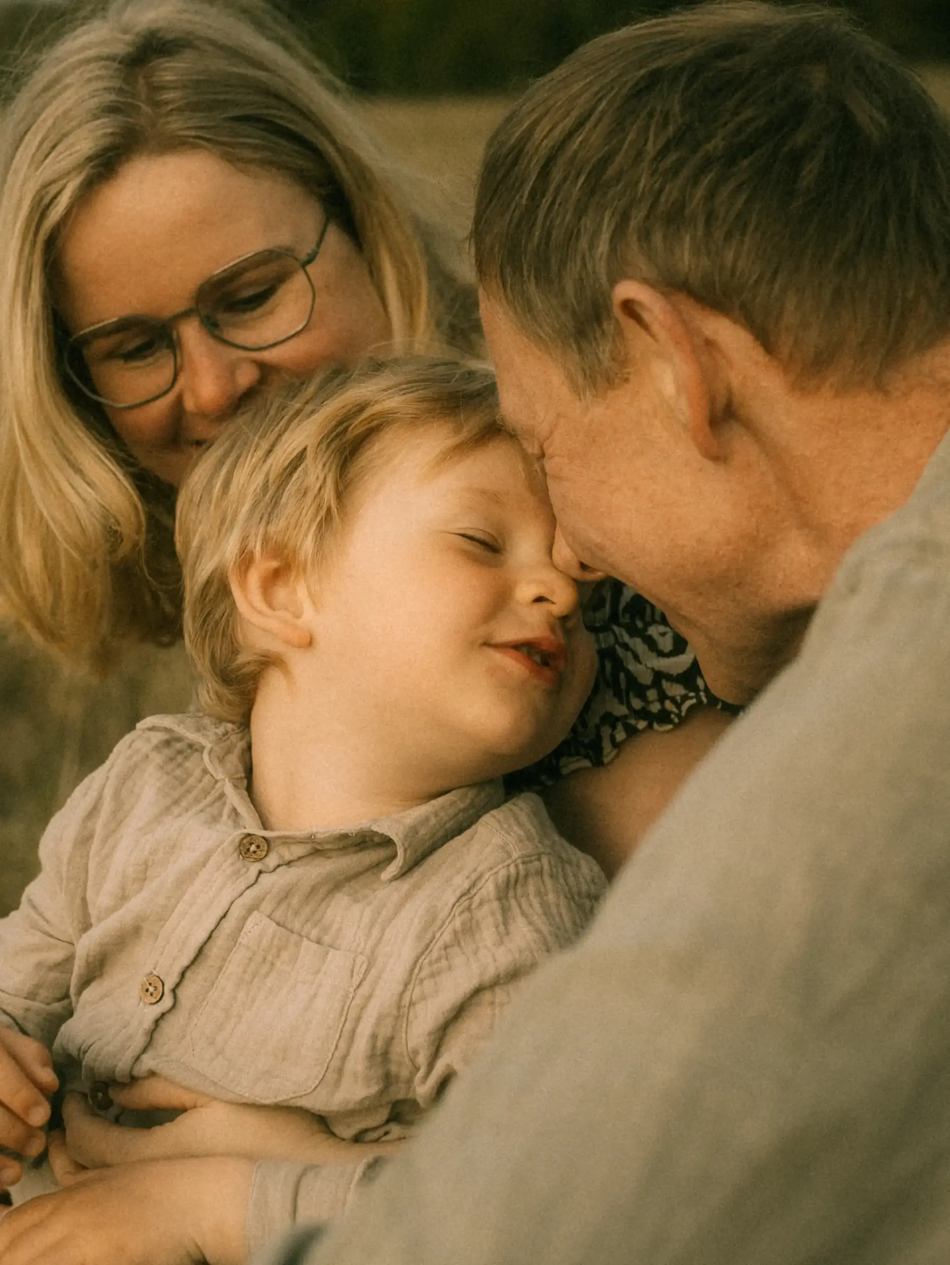 Ein Familienfotograf in Schwerin hält einen intimen Moment fest, in dem Eltern liebevoll die Stirn ihres kleinen Sohnes berühren. Das Kind lächelt glücklich in dieser warmen, natürlichen Familienaufnahme im Freien, die eine tiefe emotionale Verbundenheit ausstrahlt.