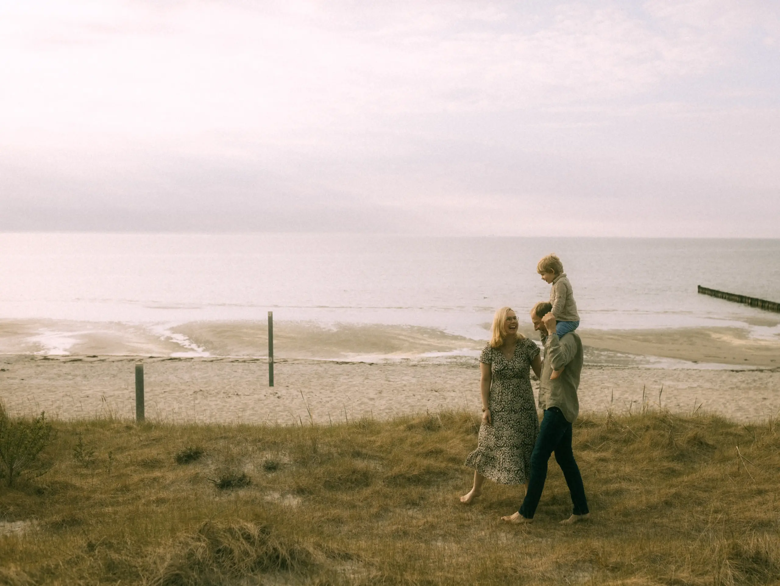 Eine glückliche Familie spaziert barfuß durch die Dünen am Strand, während der Vater sein Kind auf den Schultern trägt. Die Szene strahlt eine entspannte Atmosphäre aus und eignet sich perfekt als Inspiration für Familienfotos in Schwerin.