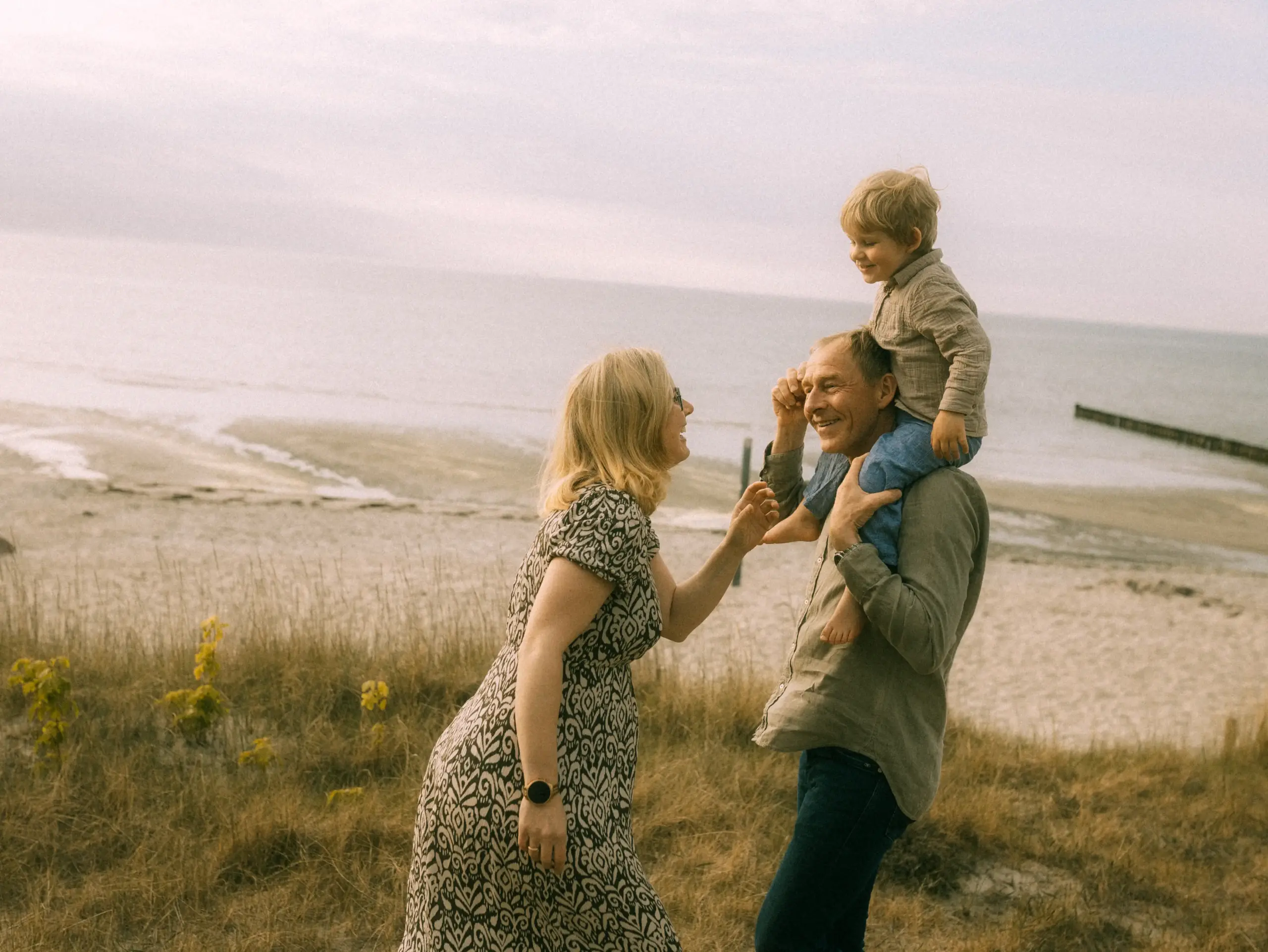 Ein glückliches Paar mit einem kleinen Kind auf den Schultern spaziert an einem sonnigen Strand in den Dünen. Die entspannte Atmosphäre fängt einen liebevollen Familienmoment ein, ideal für authentische Familienfotos in Schwerin.