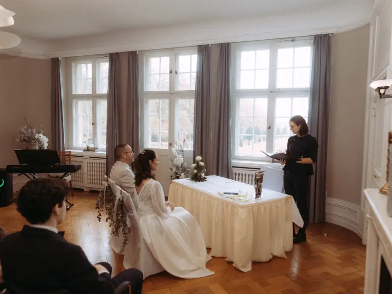 Ein Brautpaar sitzt während der standesamtlichen Trauung an einem festlich gedeckten Tisch, während eine Standesbeamtin die Zeremonie leitet. Der Raum besticht durch klassisches Interieur und große Fenster. Hochzeitsfotograf in Schwerin und MV.