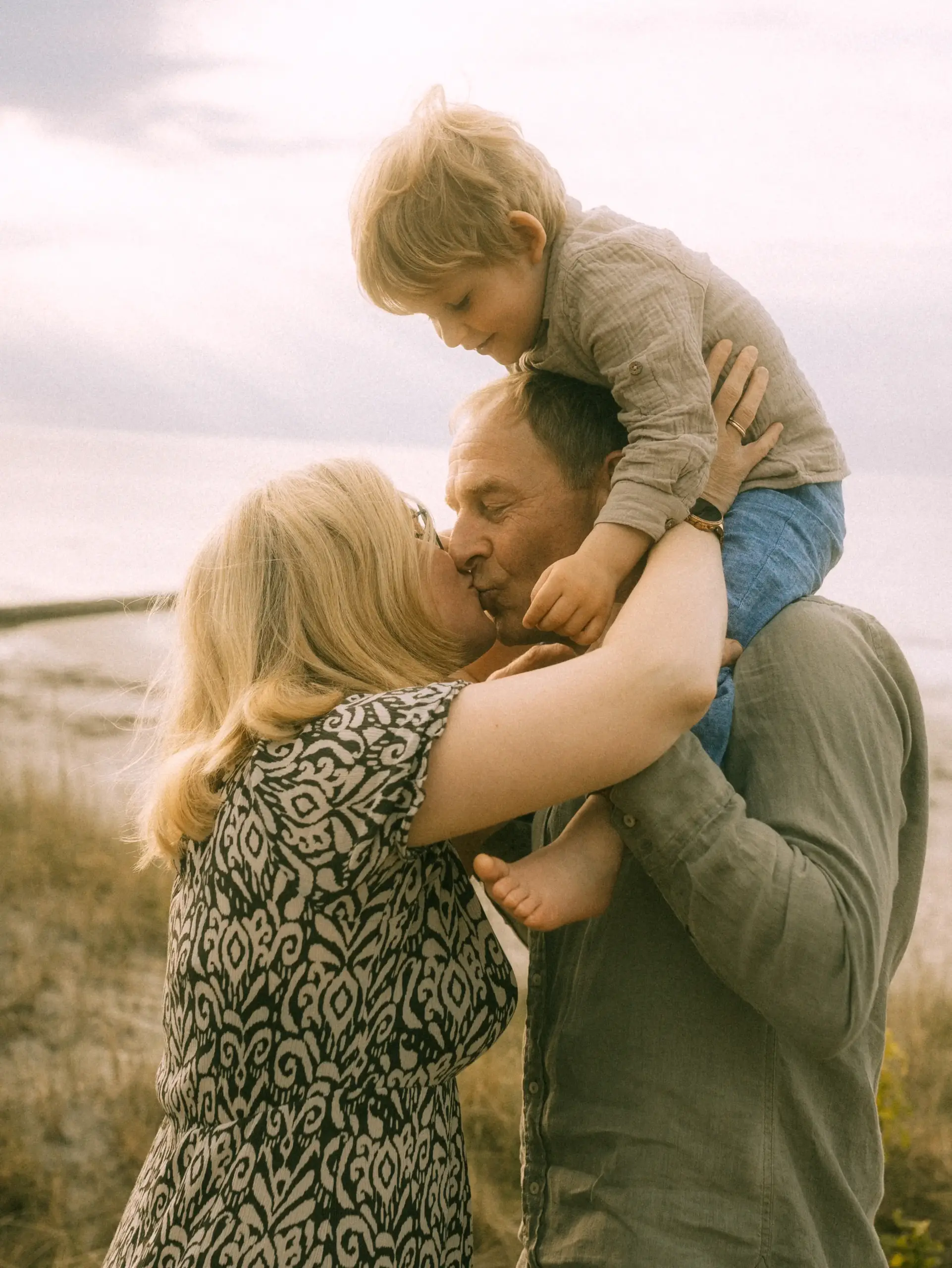 Ein Familienfotograf in Schwerin hält diesen berührenden Moment fest: Ein Paar küsst sich am Strand, während der kleine Sohn auf den Schultern des Vaters sitzt. Eine liebevolle Szene voller Verbundenheit vor einer weichen, natürlichen Küstenkulisse.