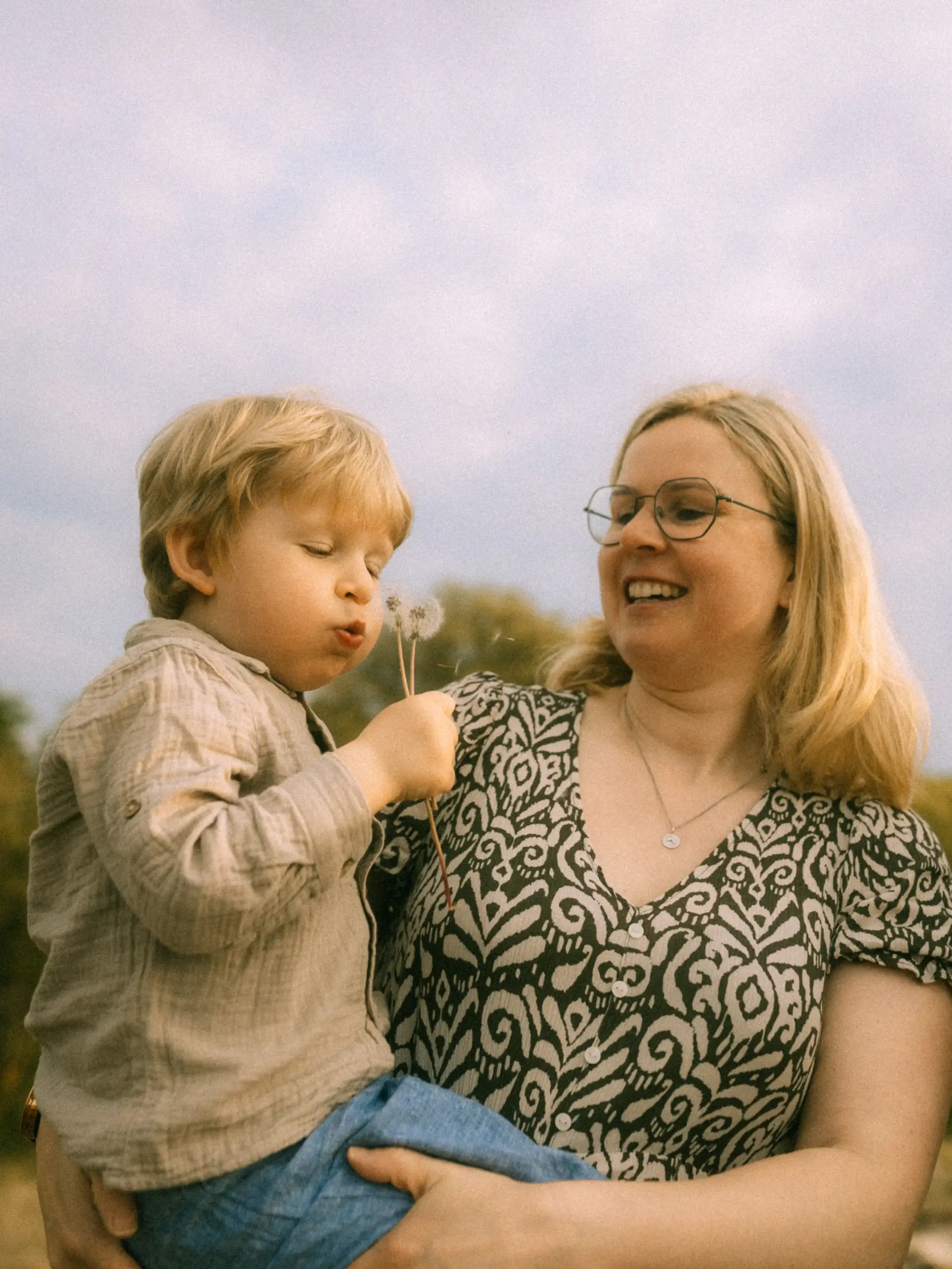 Eine lächelnde Mutter hält ihren kleinen Sohn auf dem Arm, während dieser liebevoll eine Pusteblume im Freien auspustet. Die Szene fängt einen herzlichen Moment fest, wie er für Familienfotos in Schwerin typisch ist.