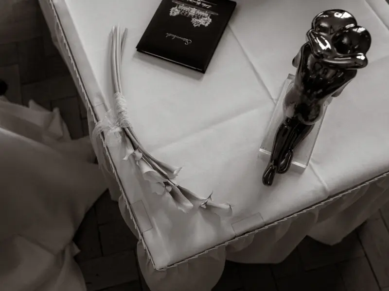 Ein elegantes Stillleben auf einem mit weißem Tuch gedeckten Tisch: Ein kunstvolles Paar-Statuette, ein weißer Callas-Brautstrauß und ein Stammbuch. Ein stimmungsvoller Moment festgehalten von Ihrem Hochzeitsfotograf in Schwerin und MV.