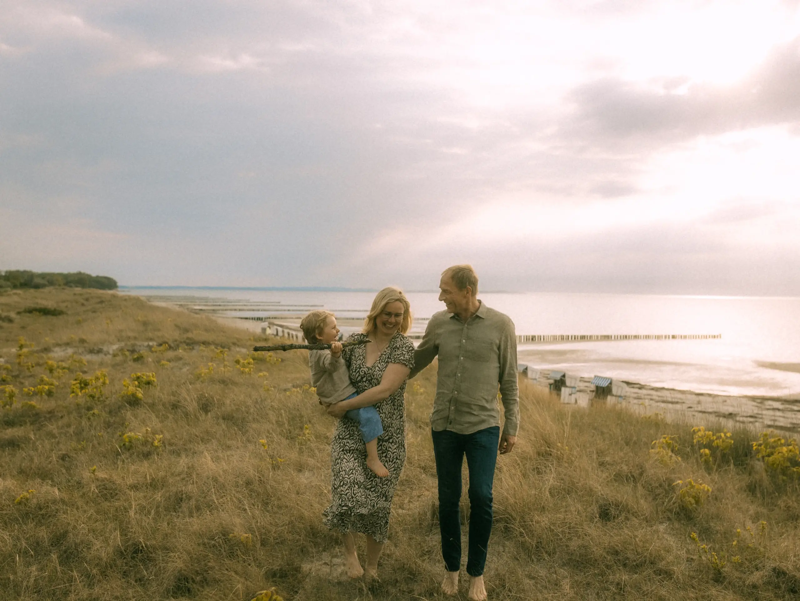 Eine glückliche Familie spaziert barfuß durch die Dünen an der Ostseeküste. Die Mutter trägt das lachende Kind auf dem Arm, während der Vater neben ihnen geht. Das stimmungsvolle Licht am Meer sorgt für eine warme Atmosphäre bei diesen natürlichen Familienfotos in Schwerin.