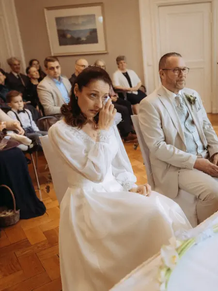Eine Braut im weißen Kleid wischt sich während der Trauung gerührt eine Träne aus dem Auge, während der Bräutigam in einem hellen Anzug neben ihr sitzt. Gäste beobachten die emotionale Zeremonie im Hintergrund. Hochzeitsfotograf in Schwerin und MV.