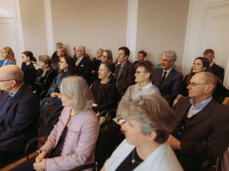 Ein aufmerksames Publikum sitzt in einem hellen, eleganten Saal bei einer Veranstaltung. Die Gäste, vorwiegend in formeller Kleidung, blicken konzentriert nach vorne. Die Szene vermittelt eine professionelle und feierliche Atmosphäre bei einer Versammlung oder Konferenz.
