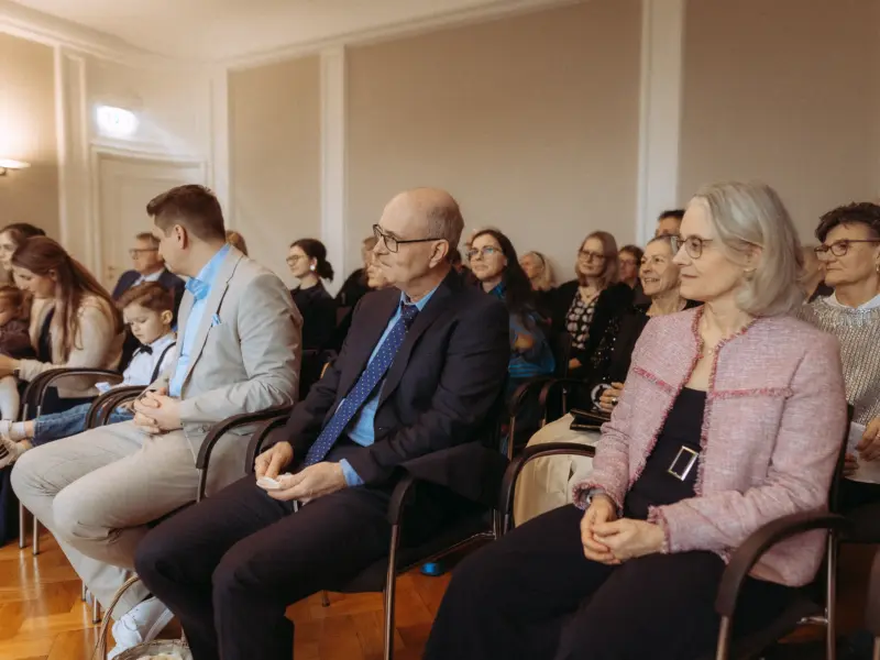 Gäste bei einer feierlichen Zeremonie sitzen in einem hellen Saal. Im Vordergrund ein Paar in eleganter Kleidung, daneben ein Mann im Anzug mit Krawatte. Das Bild wurde von einem Hochzeitsfotograf in Schwerin und MV festgehalten, der die festliche Stimmung der Gäste einfängt.