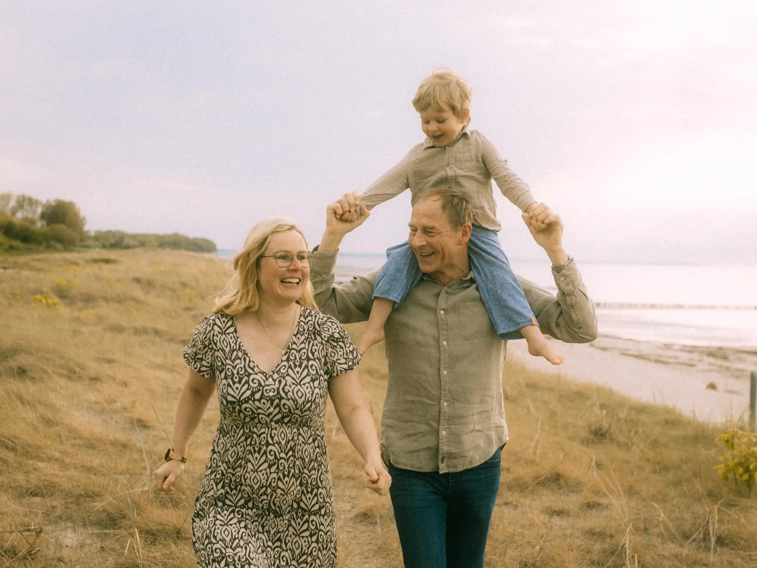 Eine glückliche Familie läuft lachend durch eine herbstliche Dünenlandschaft am Meer, während der Vater seinen kleinen Sohn auf den Schultern trägt. Authentische Familienfotos in Schwerin, die eine fröhliche Stimmung und Verbundenheit in der Natur einfangen.