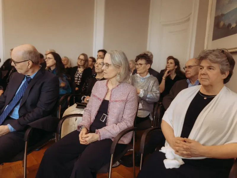 Ein aufmerksames Publikum sitzt bei einer formellen Veranstaltung in einem hellen, eleganten Saal. Im Vordergrund sind eine Frau in einem rosafarbenen Tweed-Jacket und ein Mann im dunklen Anzug zu sehen, während weitere Gäste konzentriert zuhören.