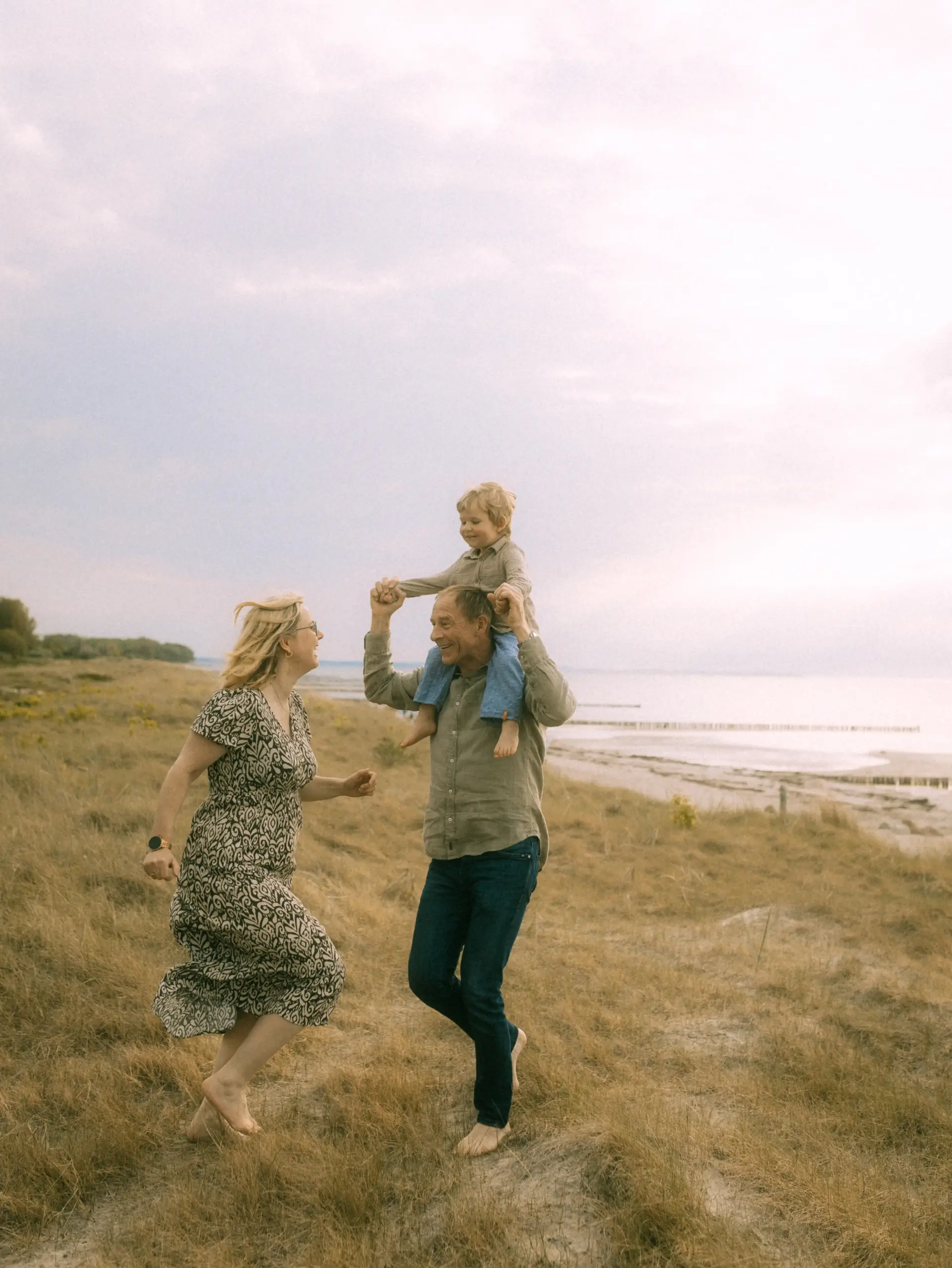 Eine glückliche Familie läuft barfuß über eine herbstliche Düne am Strand. Ein Familienfotograf in Schwerin hat den Moment festgehalten, in dem der Vater seinen Sohn auf den Schultern trägt, während die Mutter freudig nebenher läuft. Eine stimmungsvolle Aufnahme voller Lebensfreude.