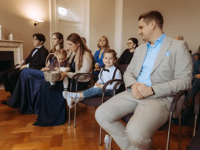 Gäste während einer feierlichen Trauung in einem eleganten Saal. Im Vordergrund sitzt ein Mann in einem hellen Anzug neben einem lächelnden Jungen mit Fliege und Hosenträgern. Eine Frau hält ein Kleinkind mit Blumenkörbchen. Ein professioneller Hochzeitsfotograf in Schwerin und MV.