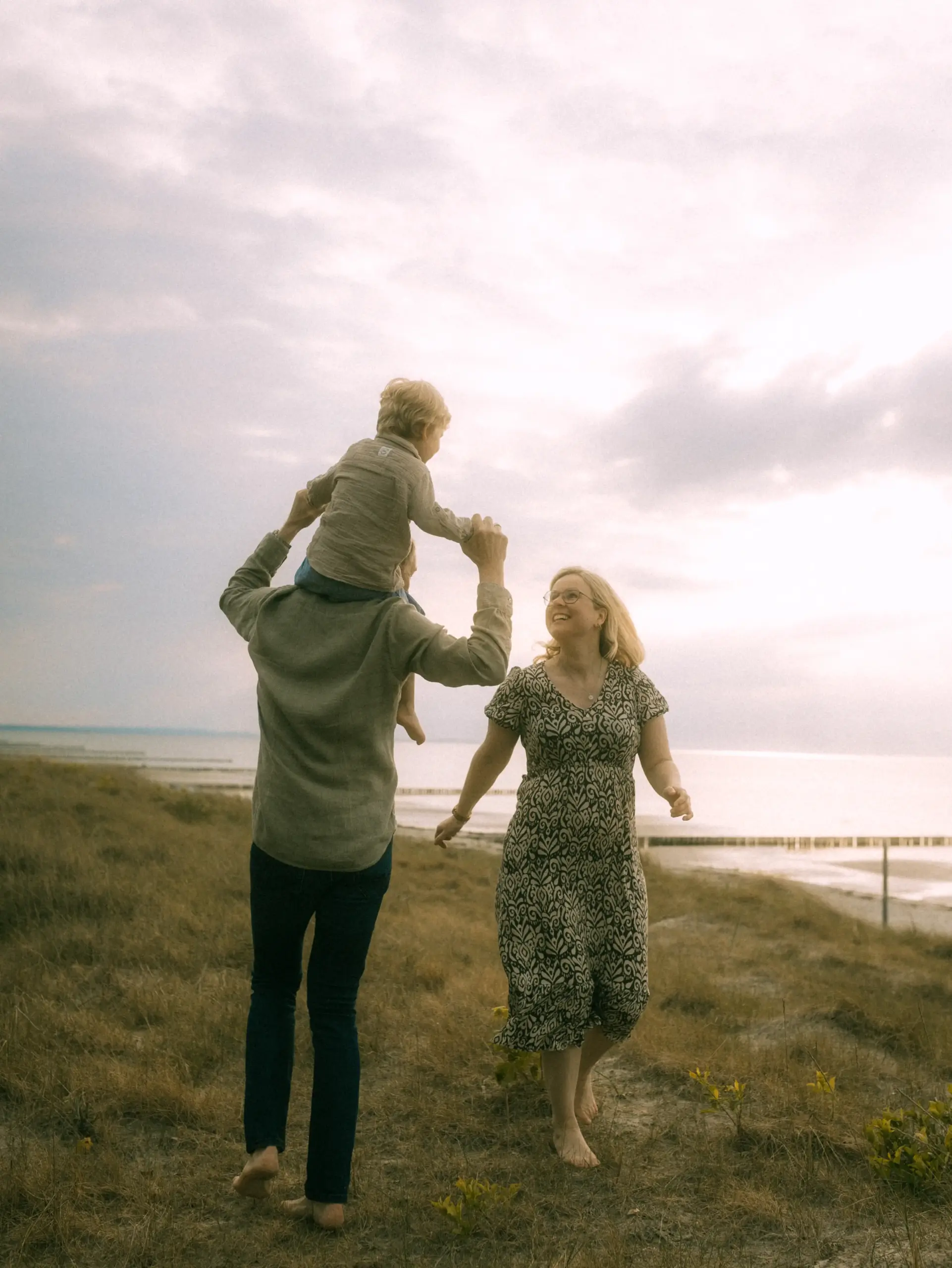 Eine glückliche Familie genießt einen Spaziergang in den Dünen am Meer: Ein Vater trägt sein Kind auf den Schultern, während die Mutter neben ihnen herläuft und lächelt. Diese authentischen Familienfotos in Schwerin halten einen unbeschwerten Moment in der Natur fest.