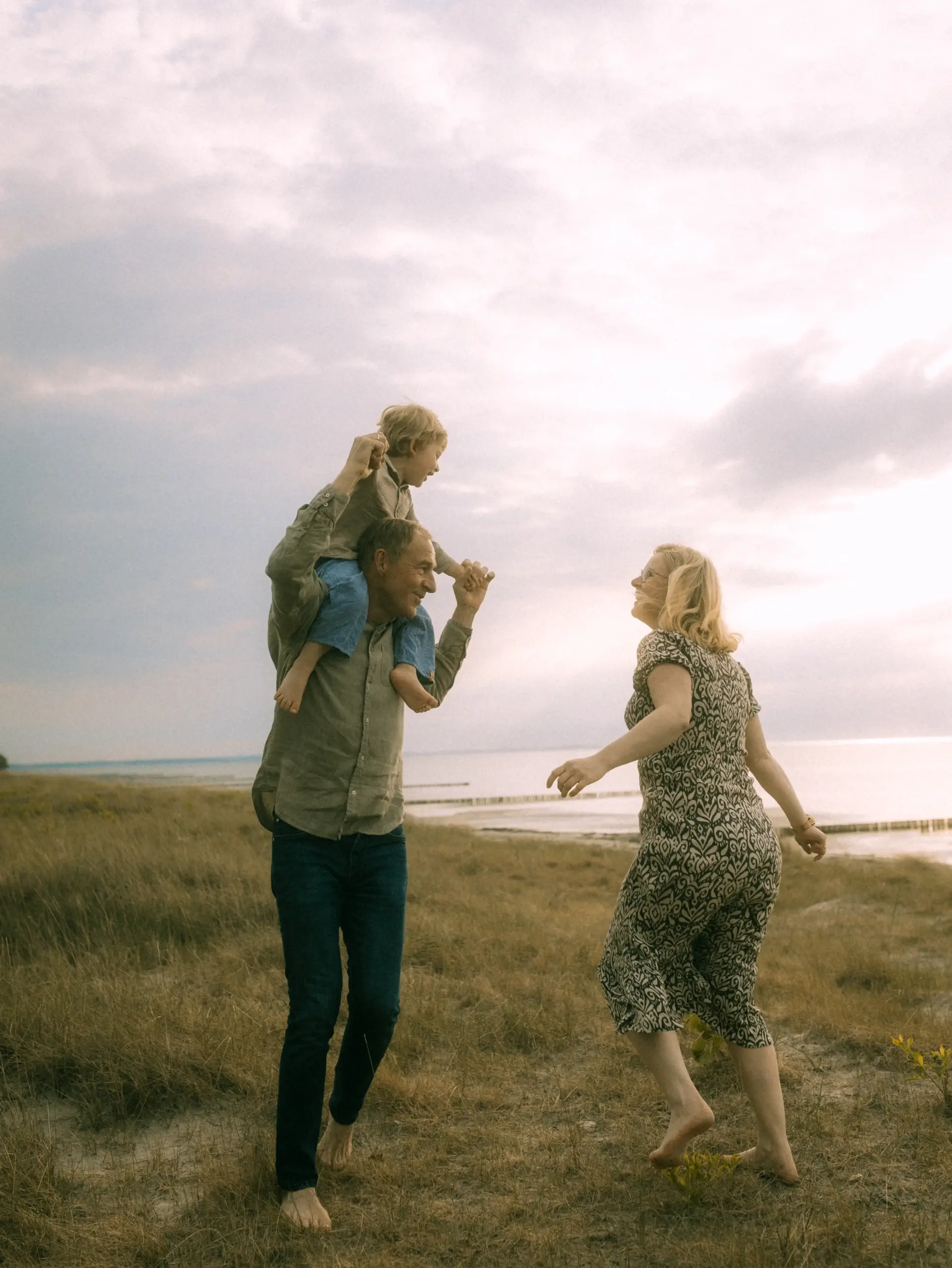 Ein glücklicher Großvater trägt seinen Enkel auf den Schultern, während die Großmutter freudig neben ihnen über eine herbstliche Küstenwiese läuft. Familienfotograf in Schwerin, der authentische und emotionale Momente in der Natur festhält.