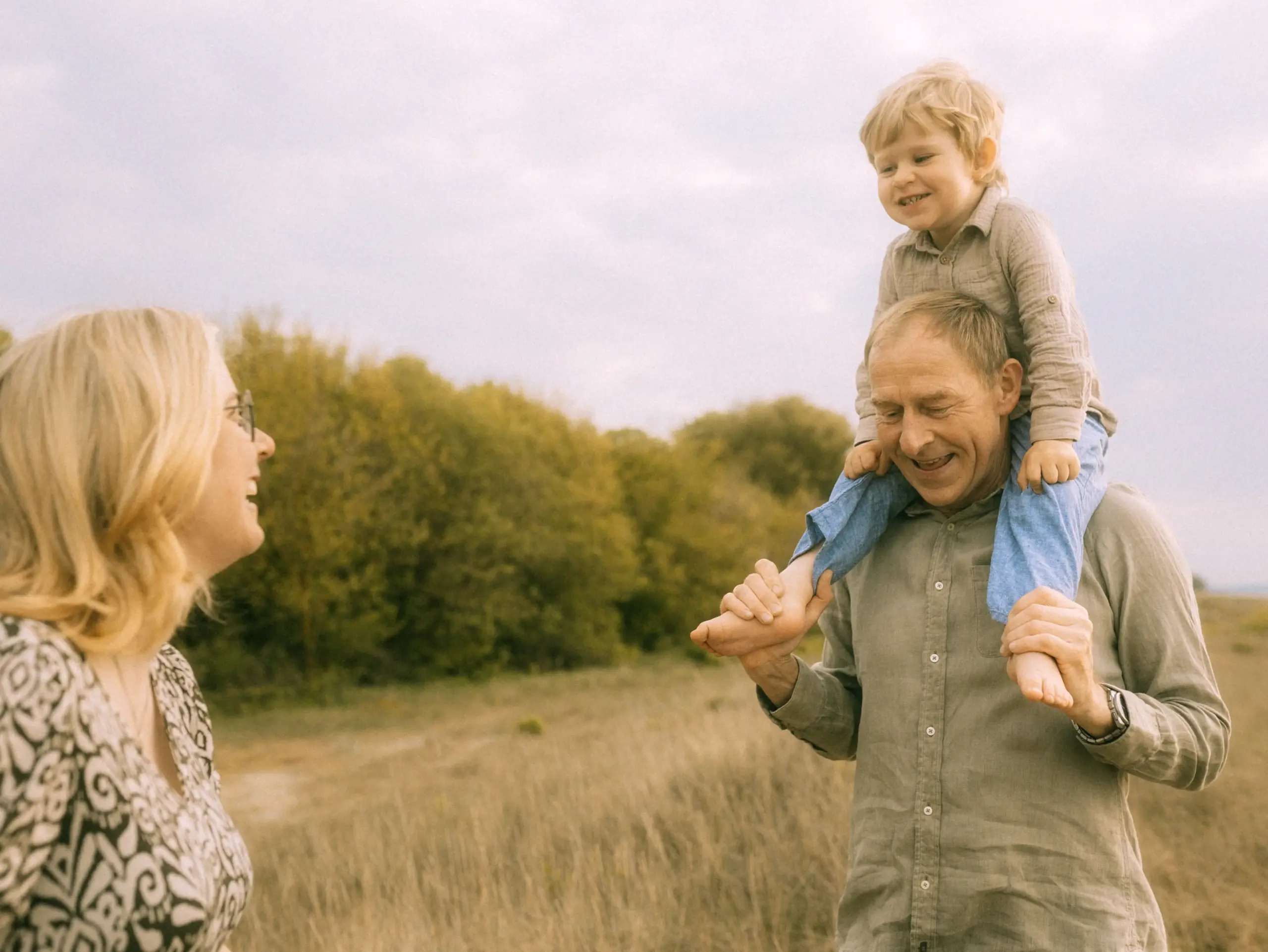 Ein glücklicher Familienfotograf in Schwerin hält einen kleinen Jungen auf den Schultern, während eine Frau lachend daneben steht. Die Familie genießt einen sonnigen Tag in der Natur, umgeben von herbstlichen Wiesen und Bäumen, was für authentische Familienfotos in Schwerin sorgt.