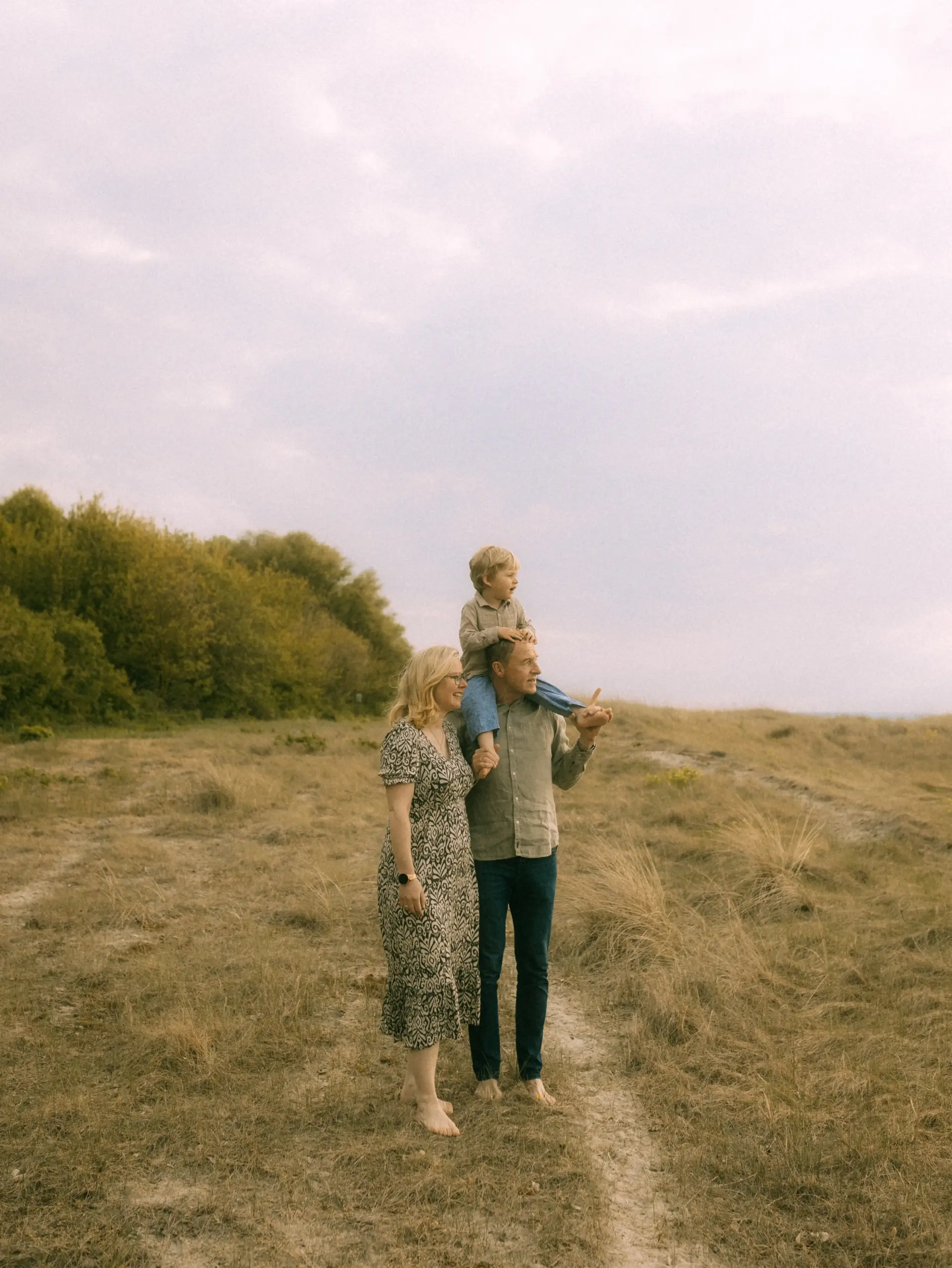 Eine junge Familie genießt einen Spaziergang in der Natur bei bewölktem Himmel. Der Vater trägt seinen Sohn auf den Schultern, während die Mutter neben ihnen geht. Authentische Familienfotos in Schwerin, die eine liebevolle Stimmung in einer herbstlichen Wiesenlandschaft einfangen.